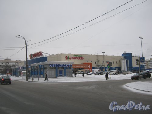 Пр. Прсвещения, дома 60. На переднем плане - автомагазин (дом 60, корпус 3), на заднем плане - гипермаркет «Карусель» (дом 60, корпус 1). Общий вид с нечётной стороны ул. Руднева. Фото 25 января 2013 г.